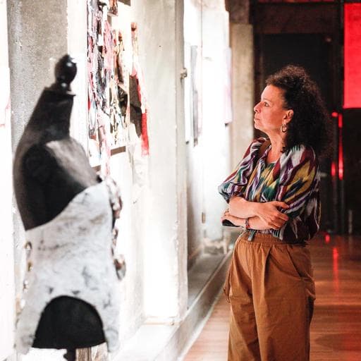 Woman with arms crossed viewing collage artwork beside a dress form in Unbound gallery.