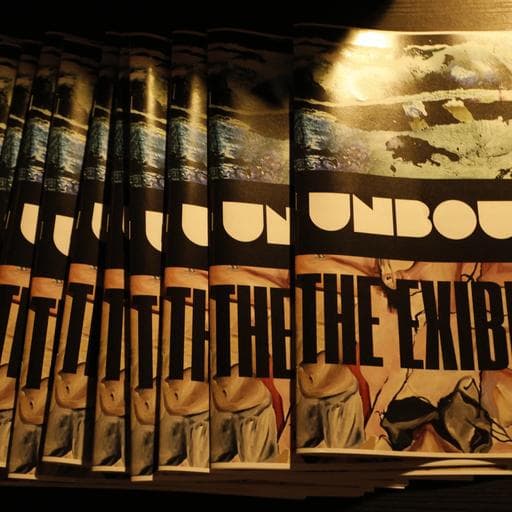 Row of Unbound exhibition catalogues reading THE EXHIBITION stacked on dark table.