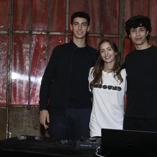 Three young people smiling behind DJ booth, one wearing white UNBOUND shirt.
