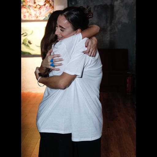 Two smiling women in white T-shirts embracing in Unbound gallery space.