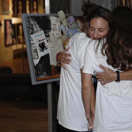 Two women hugging beside display board covered with collage posters and papers.