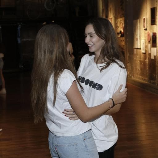 Two young women embracing in gallery hallway, one wearing UNBOUND T-shirt.