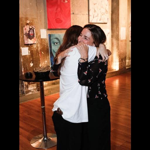 Two women embracing in gallery with colorful face paintings and wooden floor behind them.