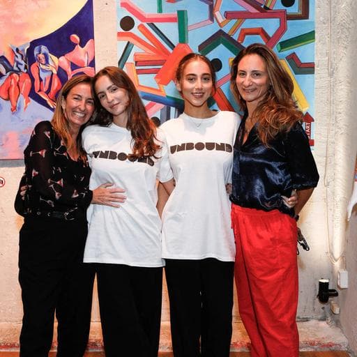 Four women posing, two wearing white UNBOUND shirts, before colorful abstract paintings.