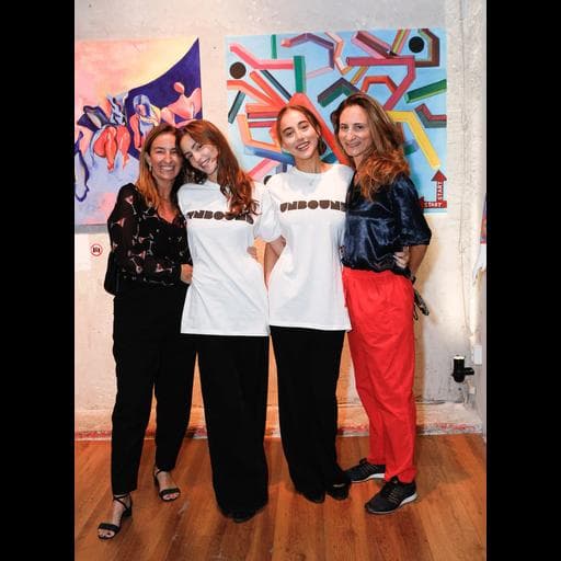 Four women standing full-length, two in UNBOUND shirts, before abstract paintings in gallery.