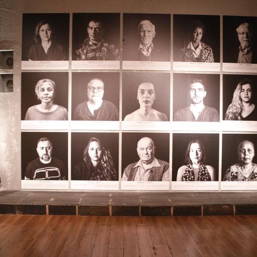 Wall of fifteen black-and-white portrait photographs in three rows above wooden floor.