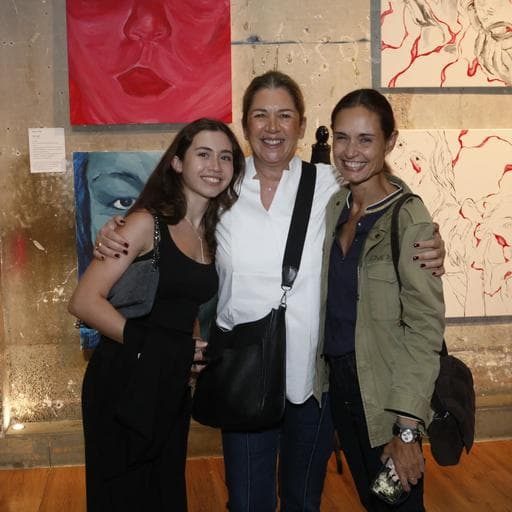 Three women posing together in front of red and blue figurative paintings at Unbound gallery.