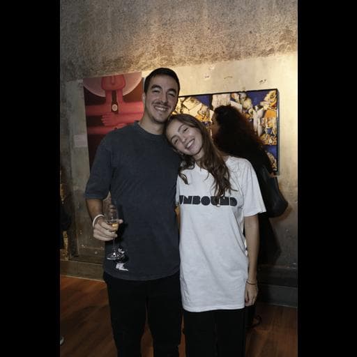 Couple posing in gallery, woman wearing white UNBOUND t-shirt, colorful paintings behind them.