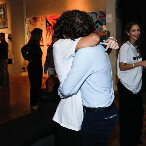 Two visitors hugging in crowded gallery with colorful paintings, woman in UNBOUND shirt nearby.