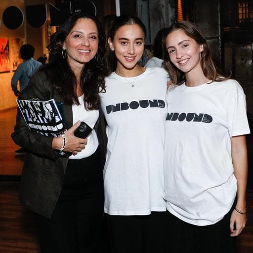 Three women posing, two wearing white UNBOUND t-shirts, one holding exhibition catalogue.