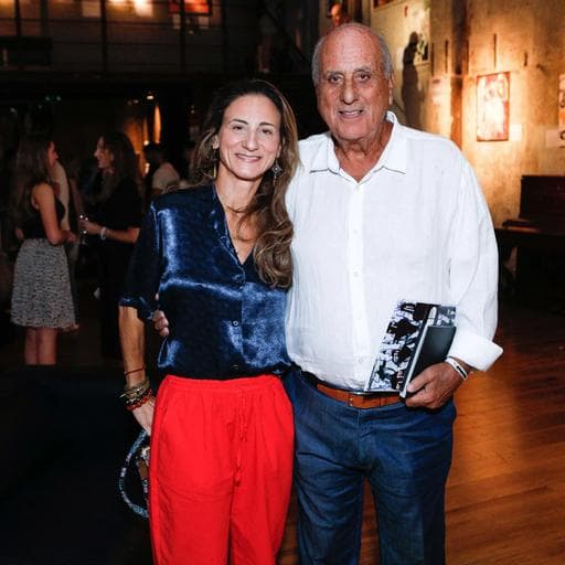 Smiling couple posing at gallery event, woman in blue blouse and red pants, crowd behind.