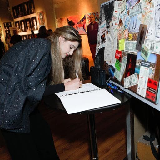 Woman signing guestbook beside collage wall covered with notes and dollar bills.