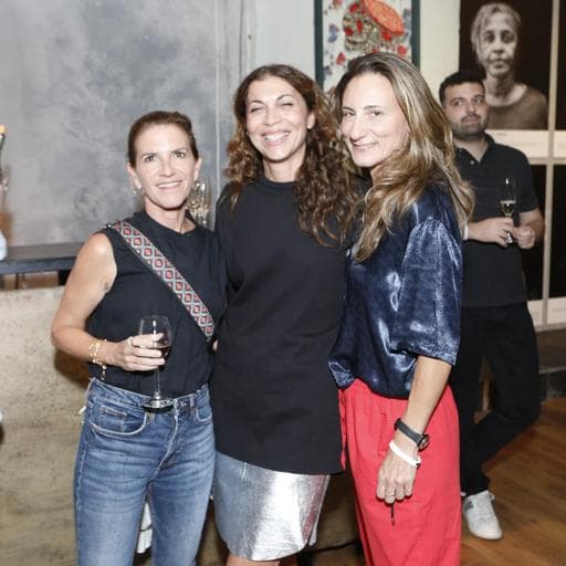 Three women smiling together holding wine glass with artworks and guests behind.