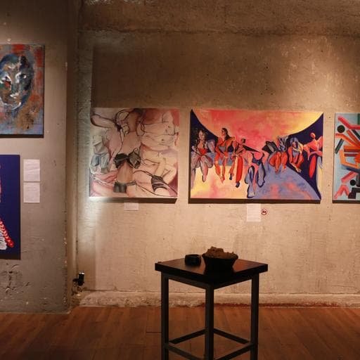 Gallery interior with colorful figurative paintings on concrete wall and small table.