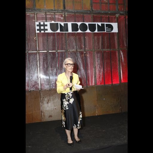 Older woman in yellow blouse and floral skirt speaking under hashtag UNBOUND sign.
