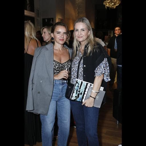Two women posing together holding drinks and catalogs at crowded gallery reception.