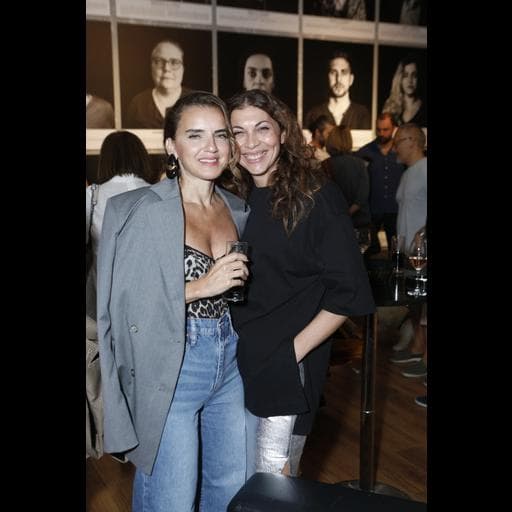 Two women posing together with drinks in front of portrait photographs at Unbound gallery.