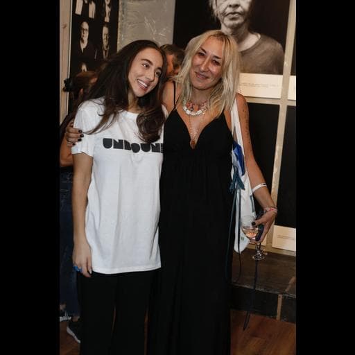 Two women posing together, one wearing a white UNBOUND t-shirt, before portrait wall.
